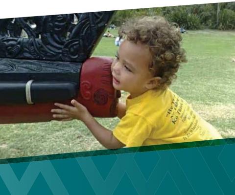 A small child with curly hair touches the front of a waka on grass.