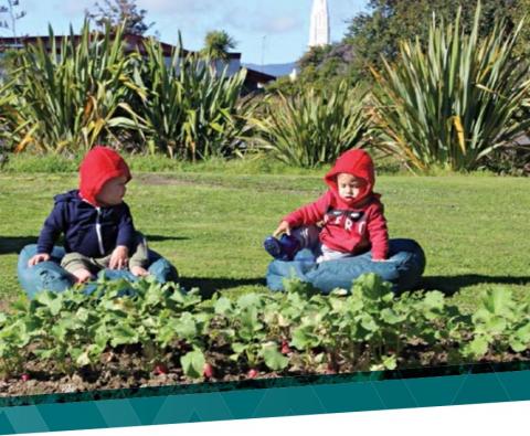 Two children wearing jackets with hoods sit on cushions on the grass next to a garden with flax in the background