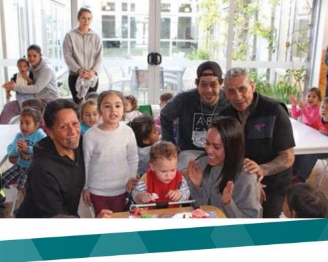 A smiling family gathers around a small child and a birthday cake with more small children sat at tables in the background.