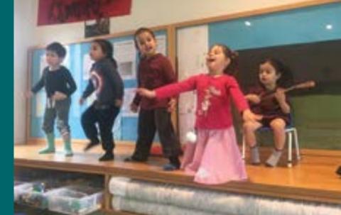 Four children stand singing on a small stage while another child sits behind with a small stringed instrument