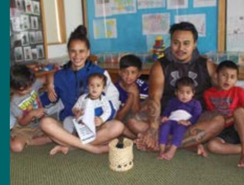 Two adults sit on the floor of a classroom with five kids of various ages sitting in close proximity